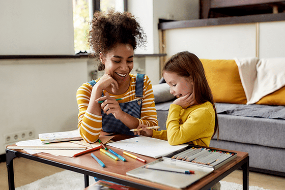 Image of a child care provider and child doing enrichment activities together in on a table in a living room next to a window
